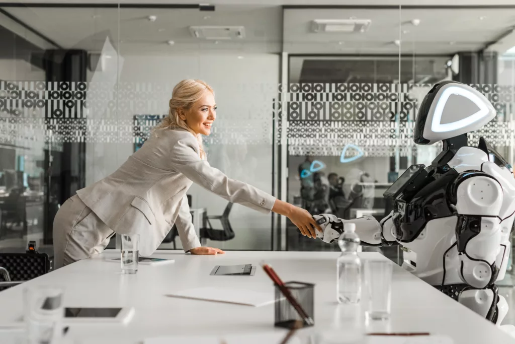 smiling businesswoman shaking hands with robot sitting at desk in meeting room